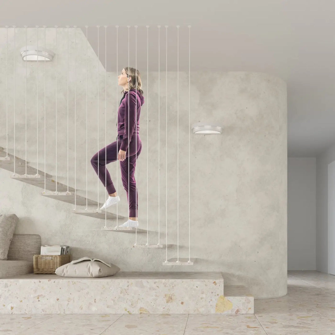 Une femme en survêtement velours violet monte un escalier minimaliste près d élégantes appliques murales helios céramique