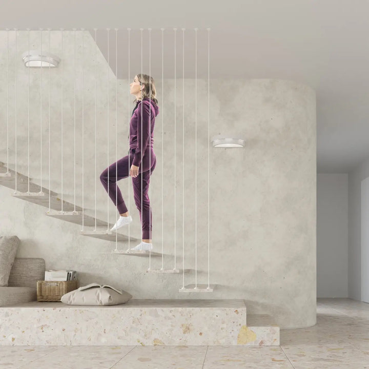 Une femme en survêtement velours violet monte un escalier minimaliste près d élégantes appliques murales helios céramique