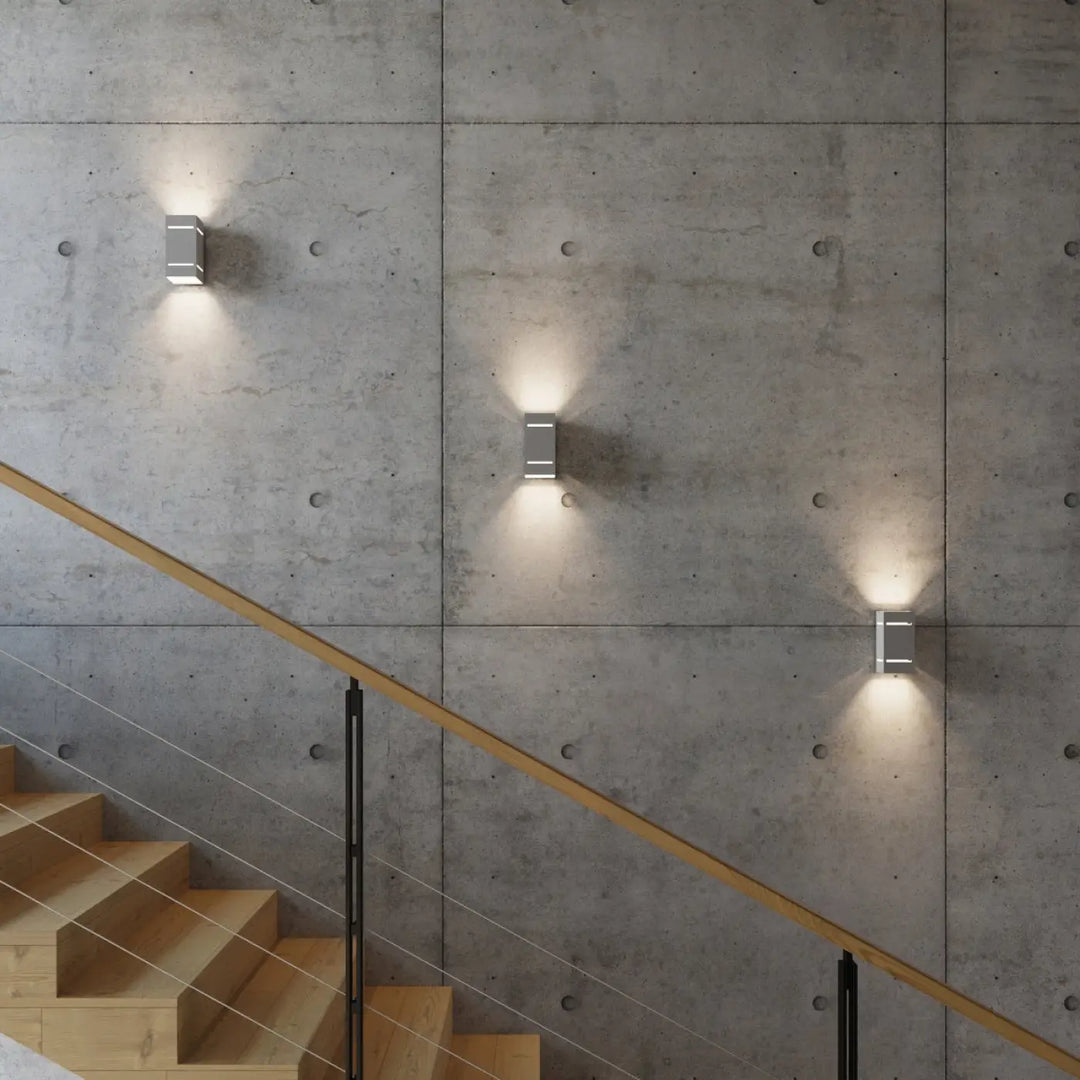 Trois appliques blocco gris en métal avec perforation linéaire projettent un spectacle lumineux sur un escalier béton