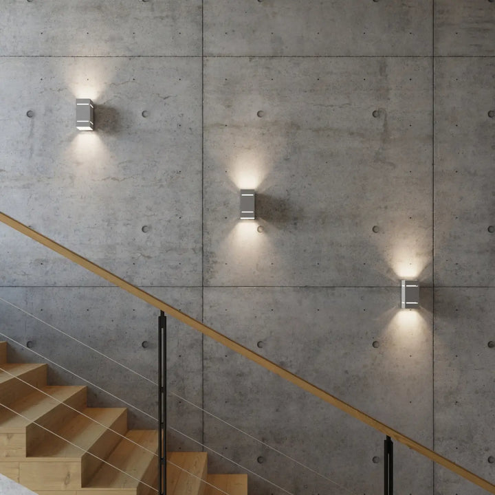 Trois appliques blocco gris en métal avec perforation linéaire projettent un spectacle lumineux sur un escalier béton