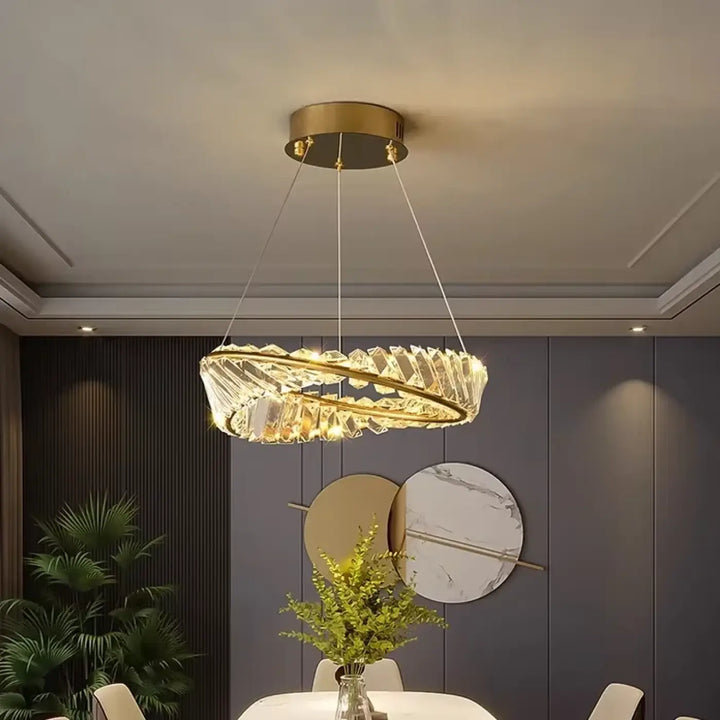 Un lustre moderne et circulaire, avec un cadre métallique aux tons dorés et des éléments transparents de type cristallin disposés en anneaux concentriques.