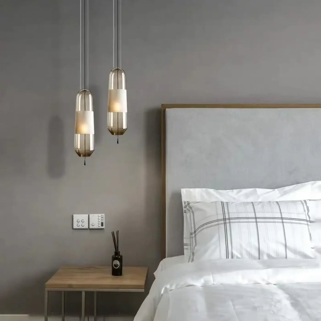 Deux suspension luminaires oxya modernes avec abat-jours en verre cylindriques et cadre métallique au-dessus d’une table de chevet