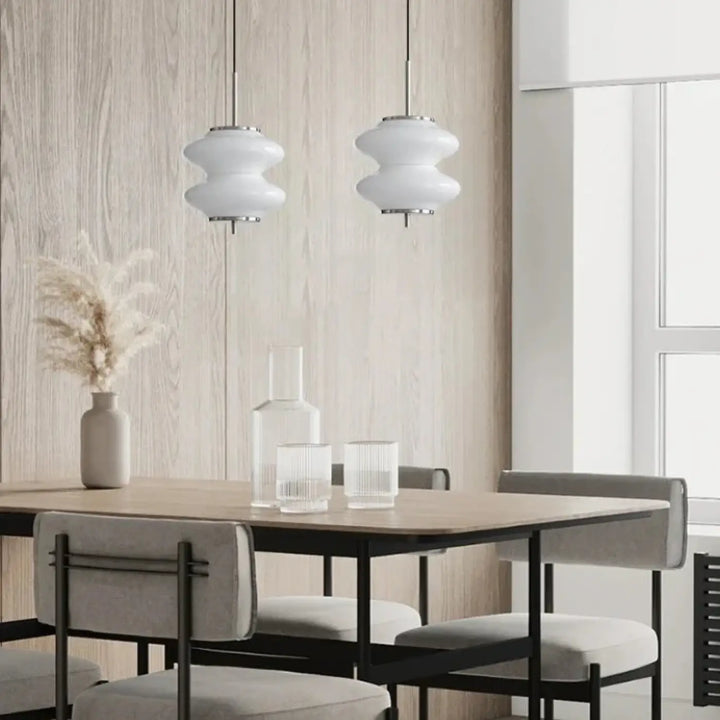 Deux suspensions luminaires en verre blanc en formes arrondies avec fixations métalliques au-dessus d’une table à manger