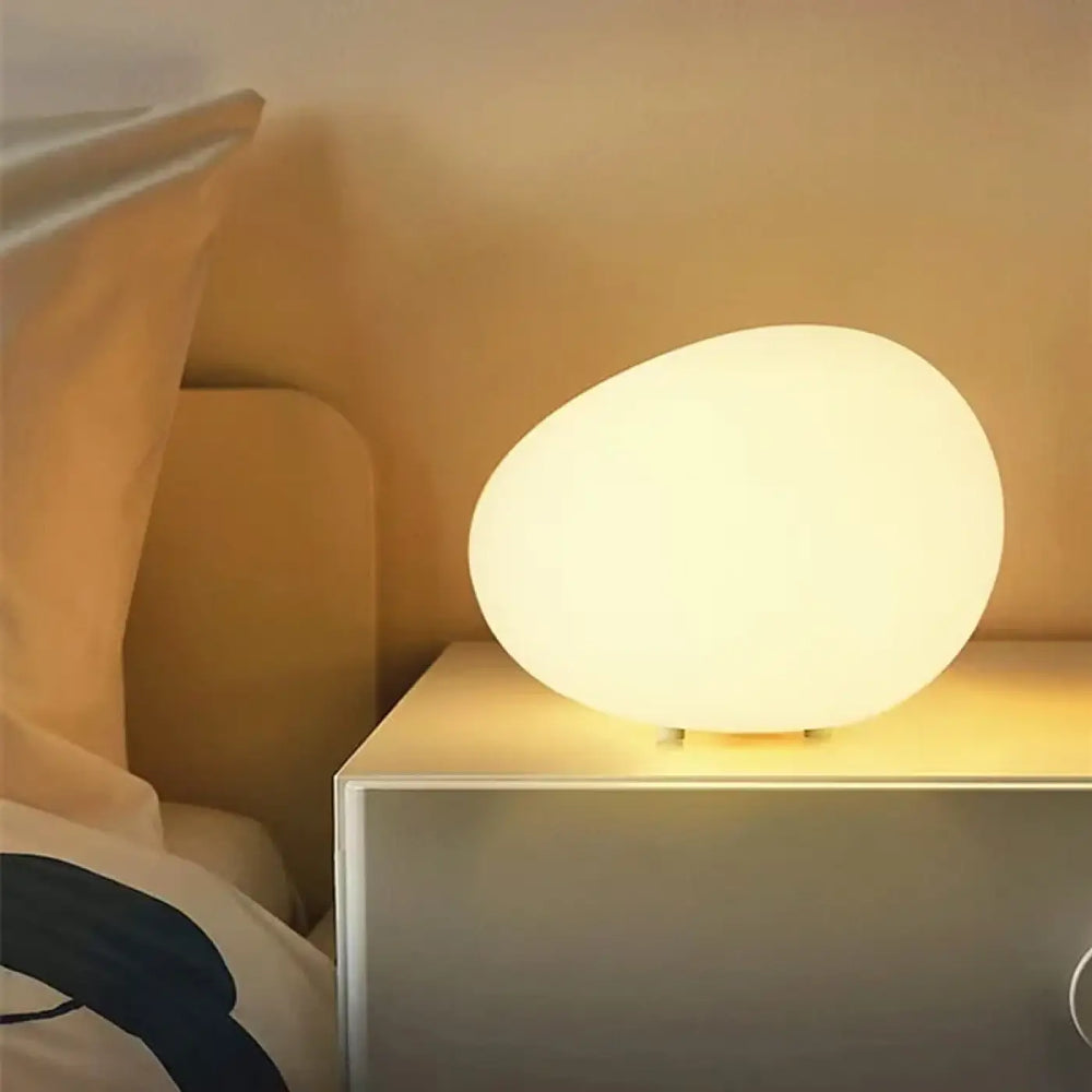 Veilleuse contemporaine inspirée d’un galet blanc doux et mat posée sur une table de chevet dans la collection eluna