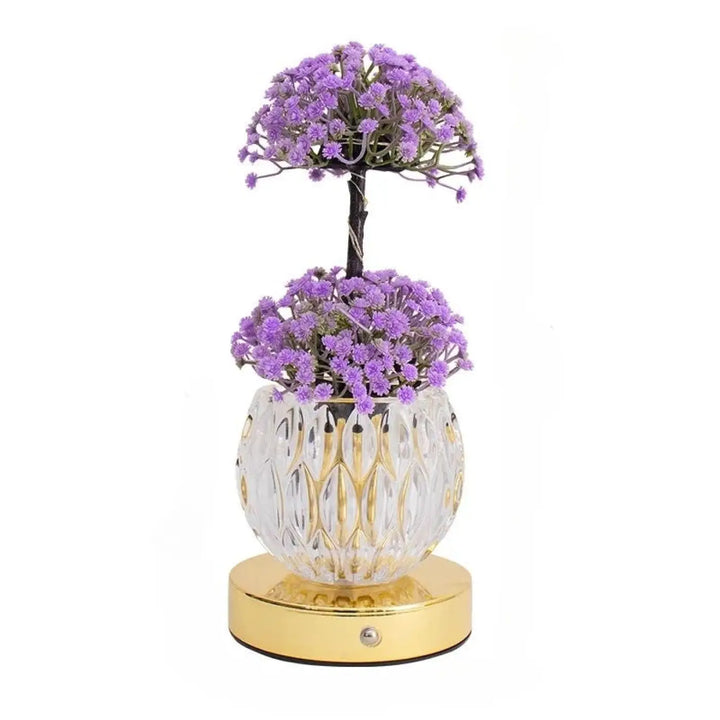 Lampe de table tulyna avec base en verre facetté clair, accents dorés, et source lumineuse ornée de fleurs violettes artificielles